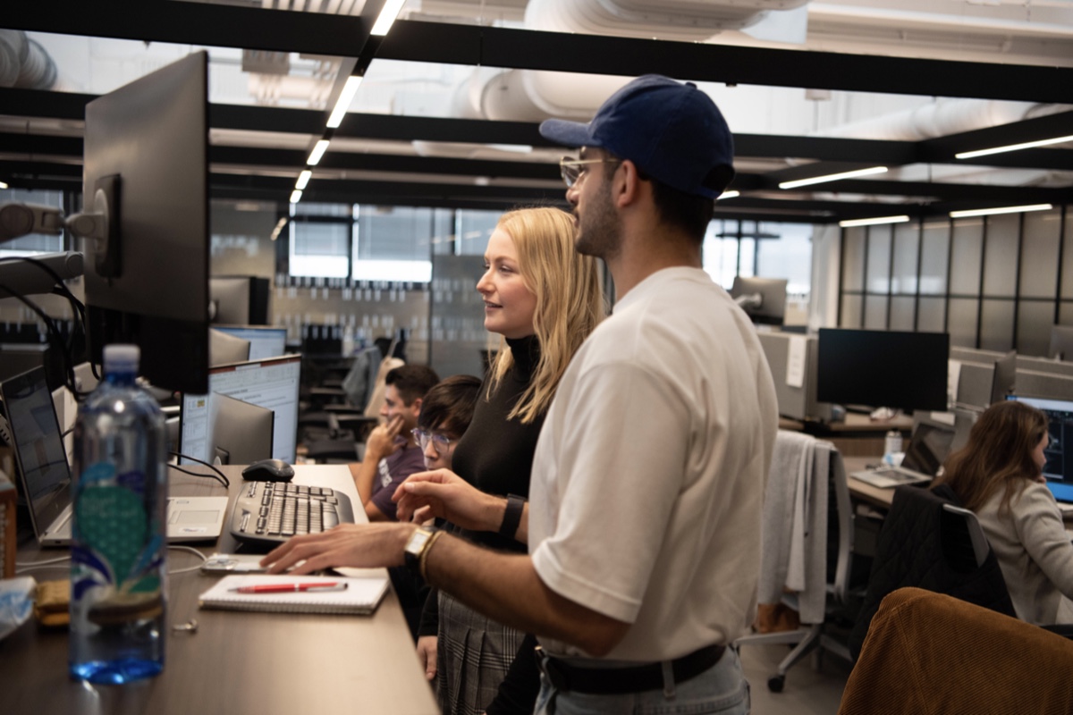 Two people are standing at a desk and looking at a computer monitor in a modern office setting. One of them is holding a pen and a notepad, while the other points at the screen. Other colleagues are working at their desks in the background.