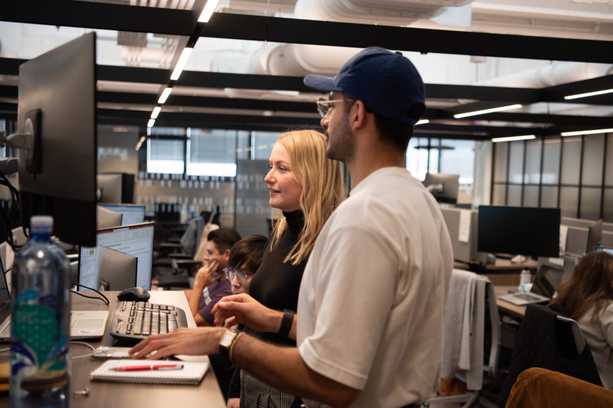 Two people stand at a desk working on a computer in an open office space. One person, wearing a black long-sleeve top, looks at the screen, while the other, in a white shirt and blue cap, points at it. Other coworkers are seated at desks in the background.