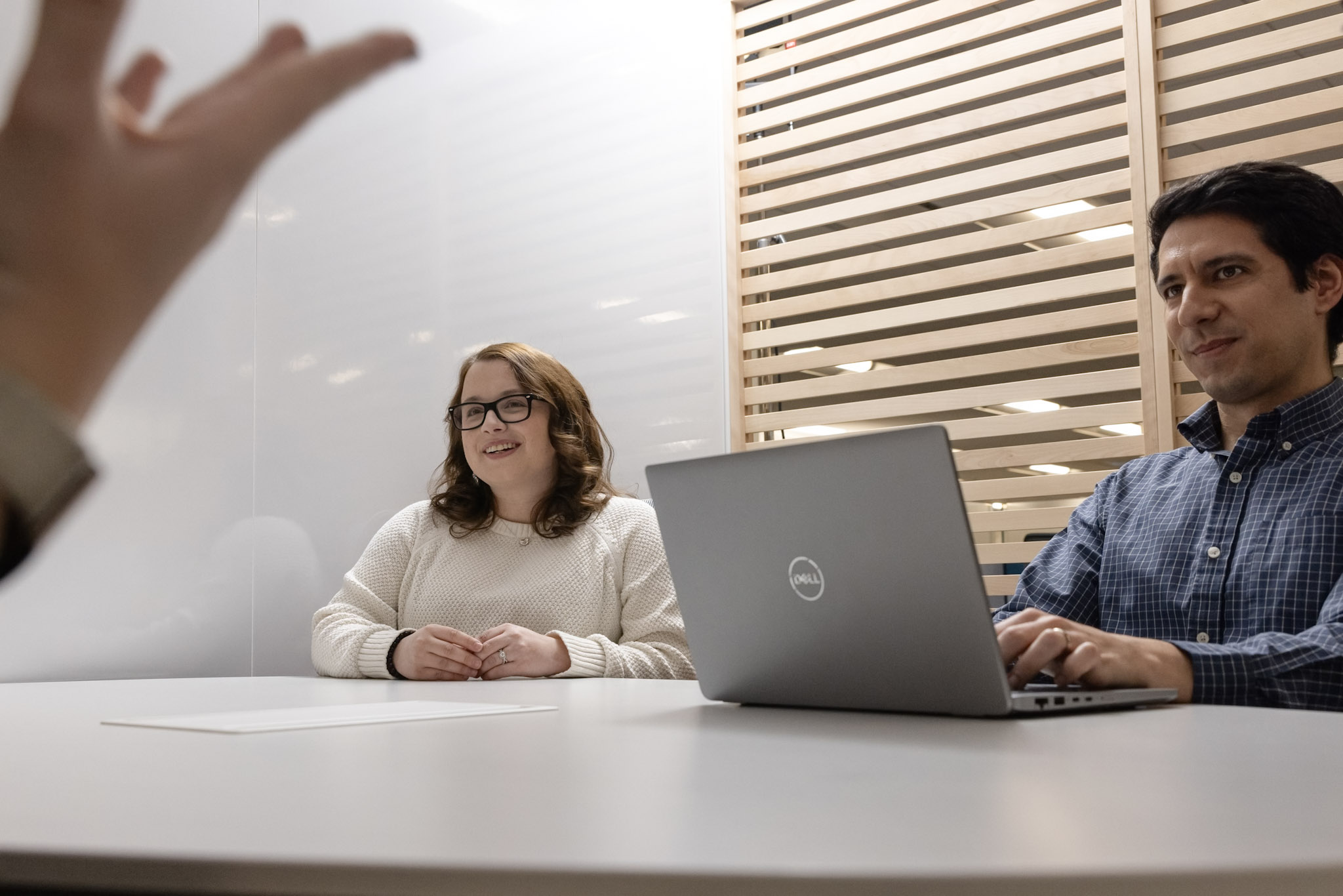 A woman and a man are sitting at a round table in an office. The woman is smiling and wearing glasses. The man is working on a Dell laptop. A hand is gesturing in the foreground. Vertical blinds partially cover the background wall.