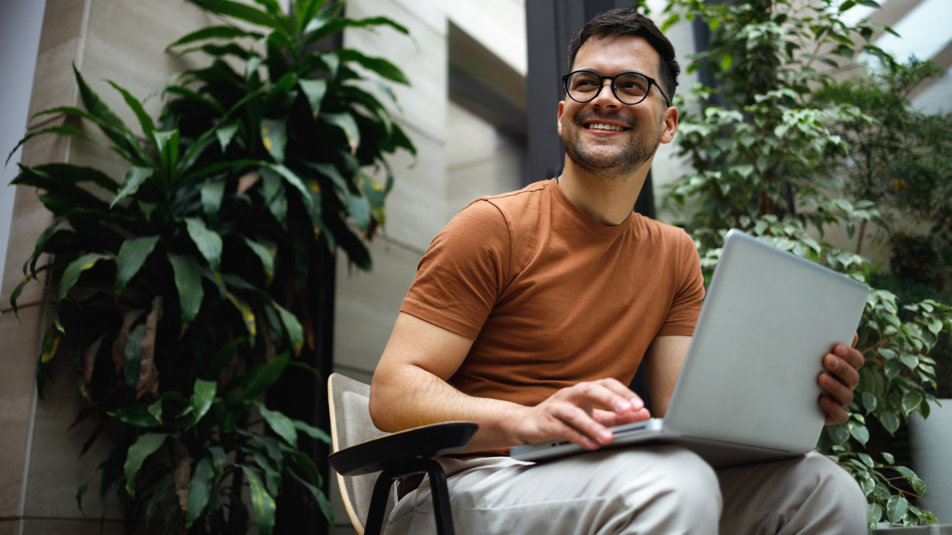 Person seated with an open laptop in a modern office environment featuring large indoor plants and natural lighting. The individual is working on the laptop while seated in a chair surrounded by greenery, representing technology, innovation and a collaborative workplace setting.