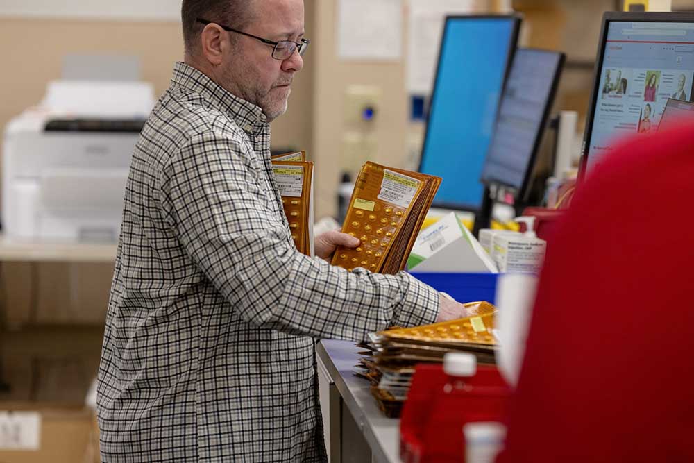 A pharmacist wearing a white lab coat and red glasses is organizing medication bottles on a shelf in a pharmacy. Another person in a white lab coat is working in the background. The shelves are filled with various pharmaceutical products.