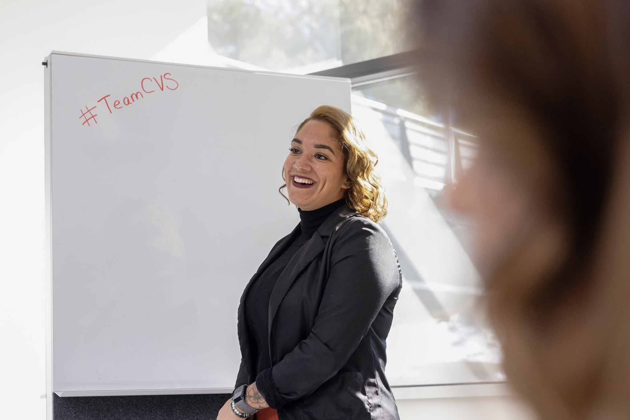 A person with shoulder-length hair is smiling and standing in front of a whiteboard. The whiteboard has “#TeamCVS” written in red marker. Sunlight is streaming through a window in the background. Part of another person’s head is visible in the foreground.