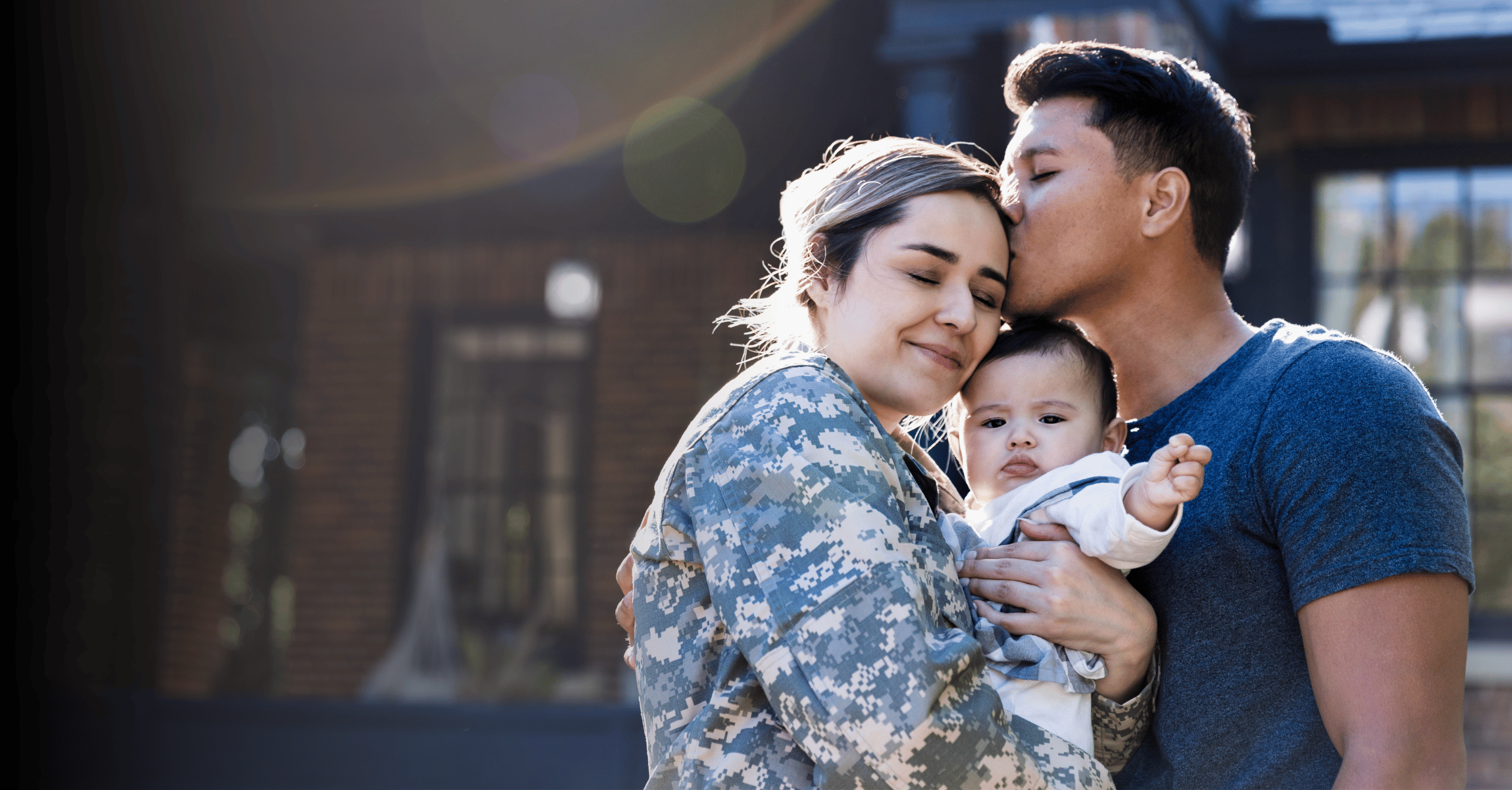 a military mom in uniform embraces her baby while a man kisses her on the forehead, capturing a tender family moment outdoors in front of a brick building.