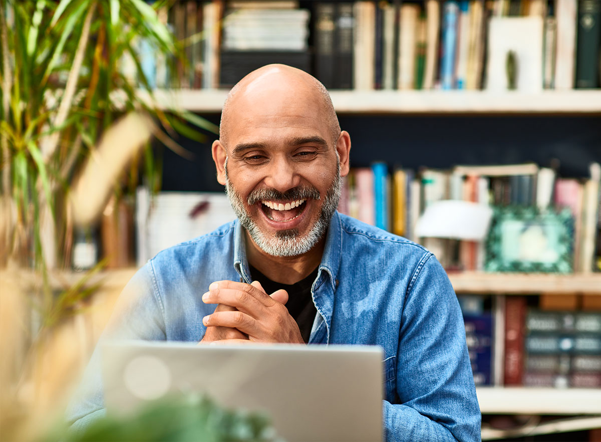 A bald man with a beard is smiling and looking at a laptop. He is wearing a blue denim shirt and sits in front of a bookshelf filled with various books. Green plants are visible in the foreground. The background is slightly blurred.