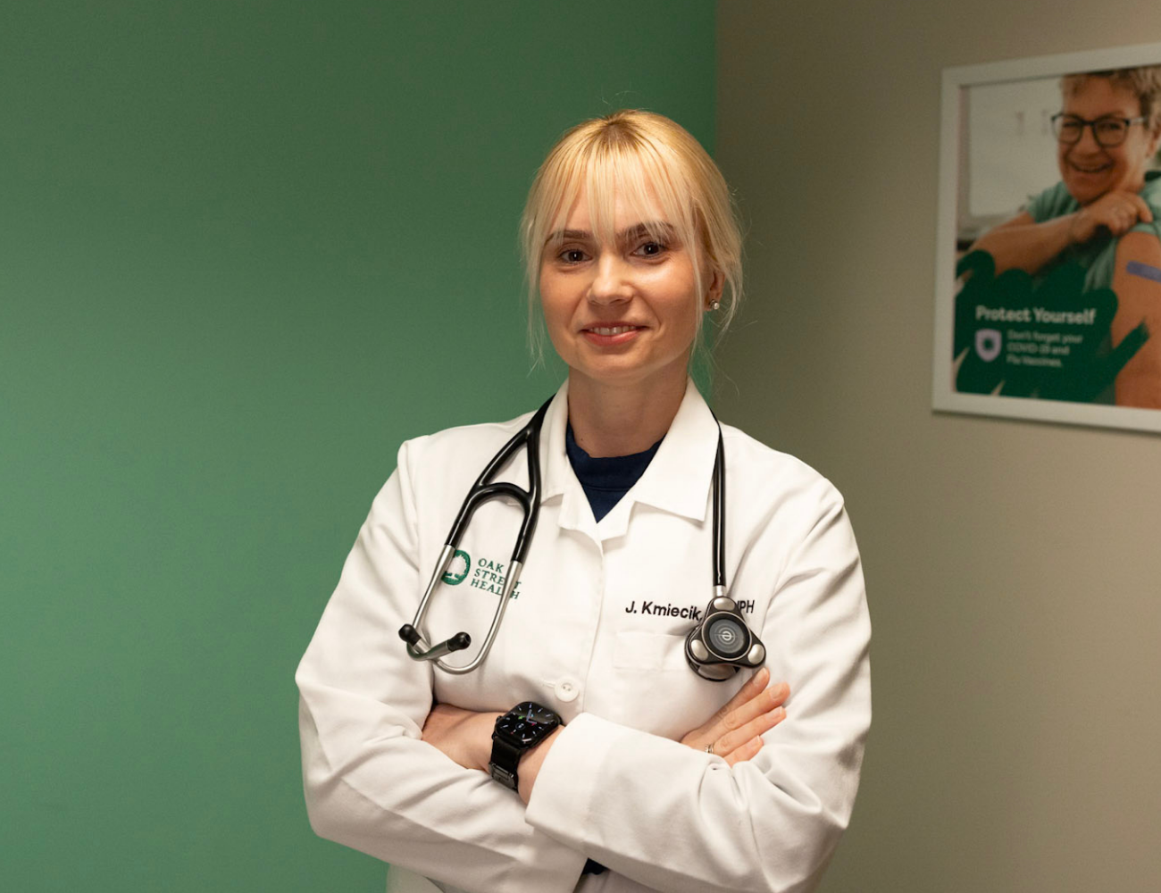 Healthcare professional wearing a white lab coat and stethoscope stands with arms crossed in a clinical exam room. A green accent wall is on the left, and a framed health awareness poster hangs on the wall in the background. Visible text on the lab coat includes “Oak Street Health.”