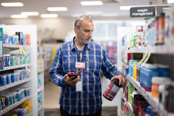 A person stands in a retail health‑care aisle, holding a smartphone in one hand and examining a large supplement container from a shelf with the other hand. Shelves on both sides are stocked with boxed and bottled products, and overhead signage reads “weight management” and “cleaning supplies.