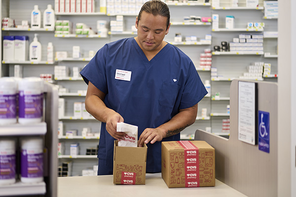 A person wearing blue scrubs and a name tag stands behind a pharmacy counter, placing a bottle of medication into a small CVS-branded cardboard box. Shelves stocked with various medications and pharmacy items are visible in the background.