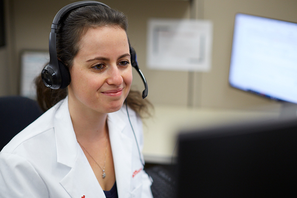 A woman with long brown hair wearing a white lab coat and a headset is looking at a computer screen. She appears to be engaged in a video call or a virtual consultation in an office setting.