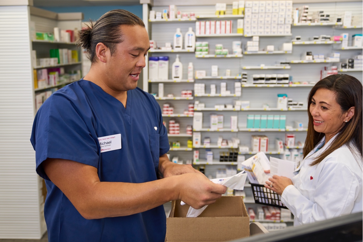 Two pharmacy professionals are standing and organizing medication in a pharmacy. The man on the left, wearing blue scrubs, opens a box, while the woman on the right, in a white lab coat, holds a medication packet. Shelves with various pharmaceutical products are in the background.