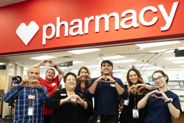 A group of people stand together inside a retail pharmacy area beneath a large red sign that reads “pharmacy” with a heart icon. Each person holds their hands in front of their chest forming a heart shape. The background shows the pharmacy counter area with overhead lighting, checkout signage, and shelving visible behind the group.