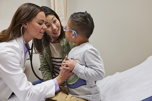 A doctor in a white coat listens to a young boy's heartbeat with a stethoscope. The boy, wearing glasses and holding his chest, sits on an examination table. A woman, likely his mother, leans in close, smiling at her son.