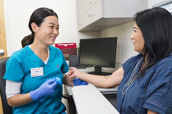 A healthcare professional in teal scrubs and gloves is smiling while holding the hand of a patient sitting in a clinic. The patient is seated in a striped top. A computer and medical supplies are visible in the background.