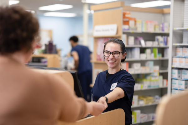 A pharmacy tech stands behind a counter, reaching out to exchange an item with a customer, with medication shelves and another staff member visible in the background.