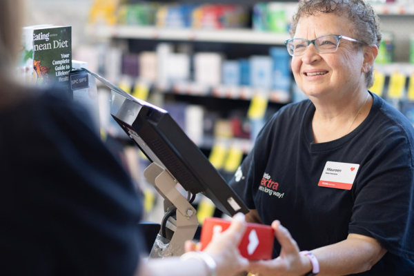 Maurenn, a store employee stands at a checkout counter, holding a small red item toward a customer, with shelves of products visible in the background.