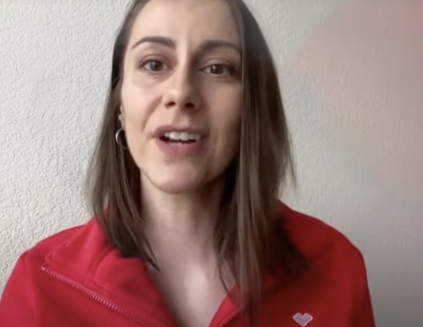 A person with straight, shoulder-length brown hair is wearing a red zip-up sweater with a small white heart on it. They are standing in front of a plain, light-colored wall, looking at the camera with a neutral expression.