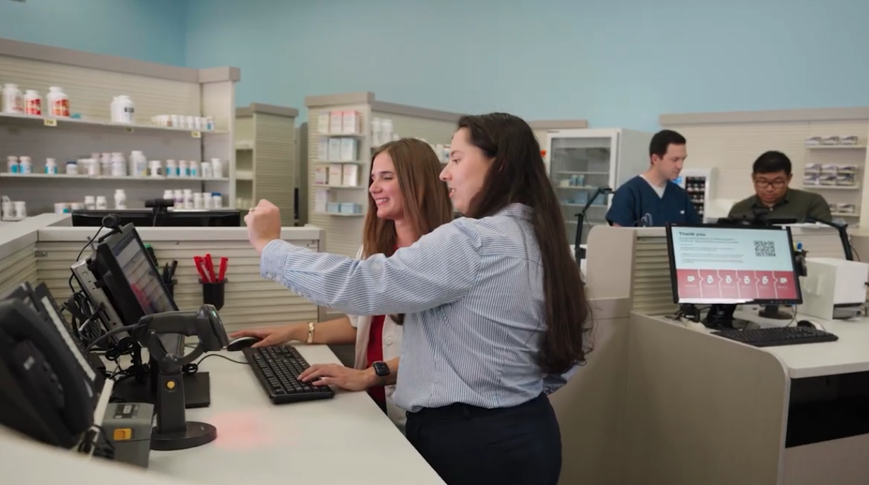 Two employees work together at a pharmacy workstation, reviewing information on a computer monitor at the prescription counter. Shelves stocked with medication containers line the background, while additional staff members work at nearby stations. The pharmacy is brightly lit with computer equipment, scanners, and customer service terminals visible throughout the workspace.