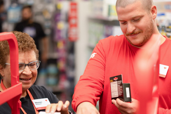 Two people stand at a service counter inside a store. One person wearing a red long‑sleeve shirt with a CVS logo holds a small boxed product, while the other person with short curly hair and glasses gestures toward the counter. Store shelving and products are visible in the background, with red checkout fixtures partially framing the foreground.