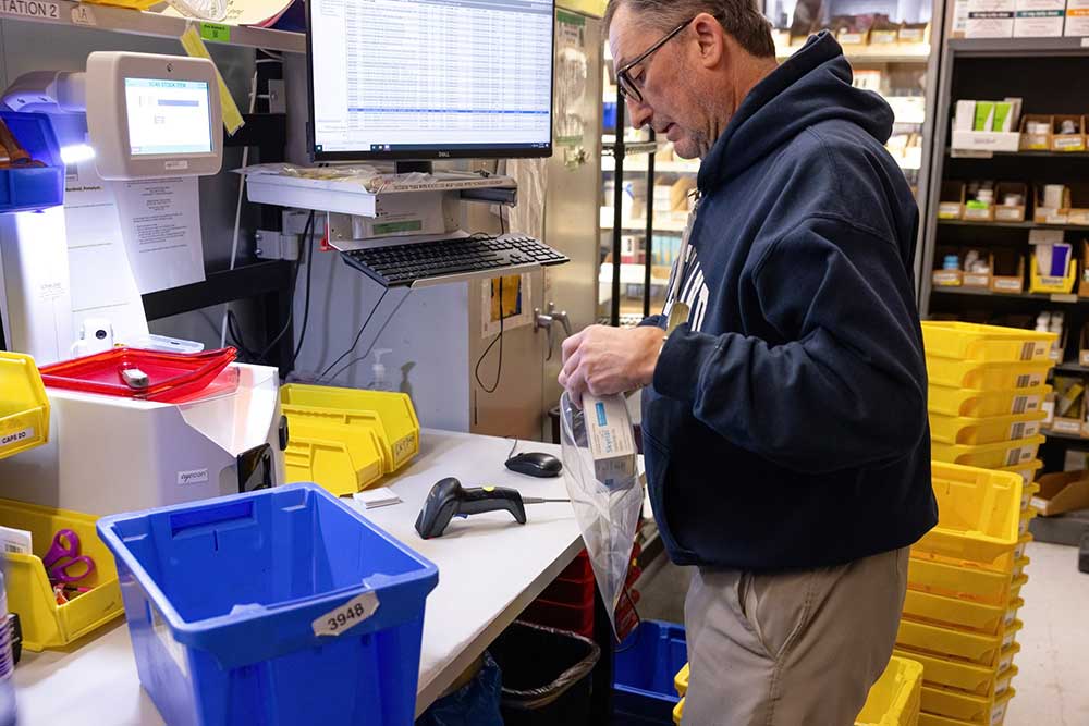 A healthcare worker, wearing a maroon scrub top and an ID badge on a red lanyard, holds up and inspects an IV bag in a medical supply room with metal shelves and white boxes.