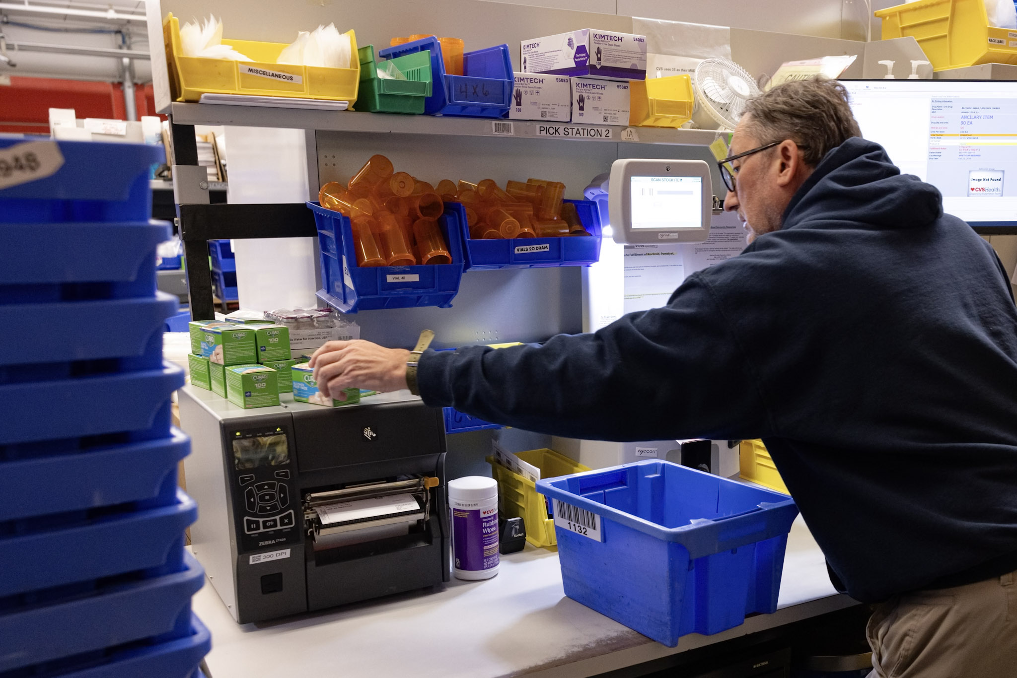 A pharmacist in a white coat handing a prescription bottle to a smiling man in blue scrubs inside a pharmacy. Shelves filled with medication bottles are visible in the background.