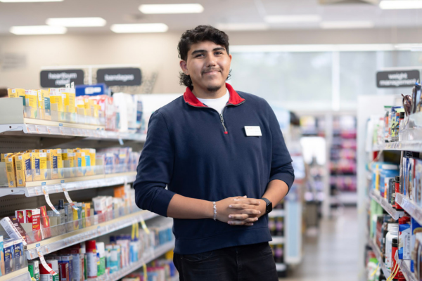 A person stands in the middle of a brightly lit retail aisle with shelves of pharmacy products on both sides. The individual is wearing a dark long-sleeve top with a visible name badge and has hands clasped at waist level, while boxed health and personal care items line the shelves in the foreground and background.