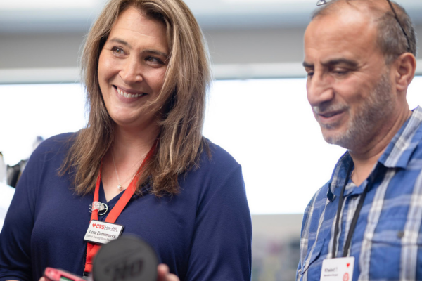 Two store employees stand indoors reviewing an item together, with shelving and store fixtures visible in the background.