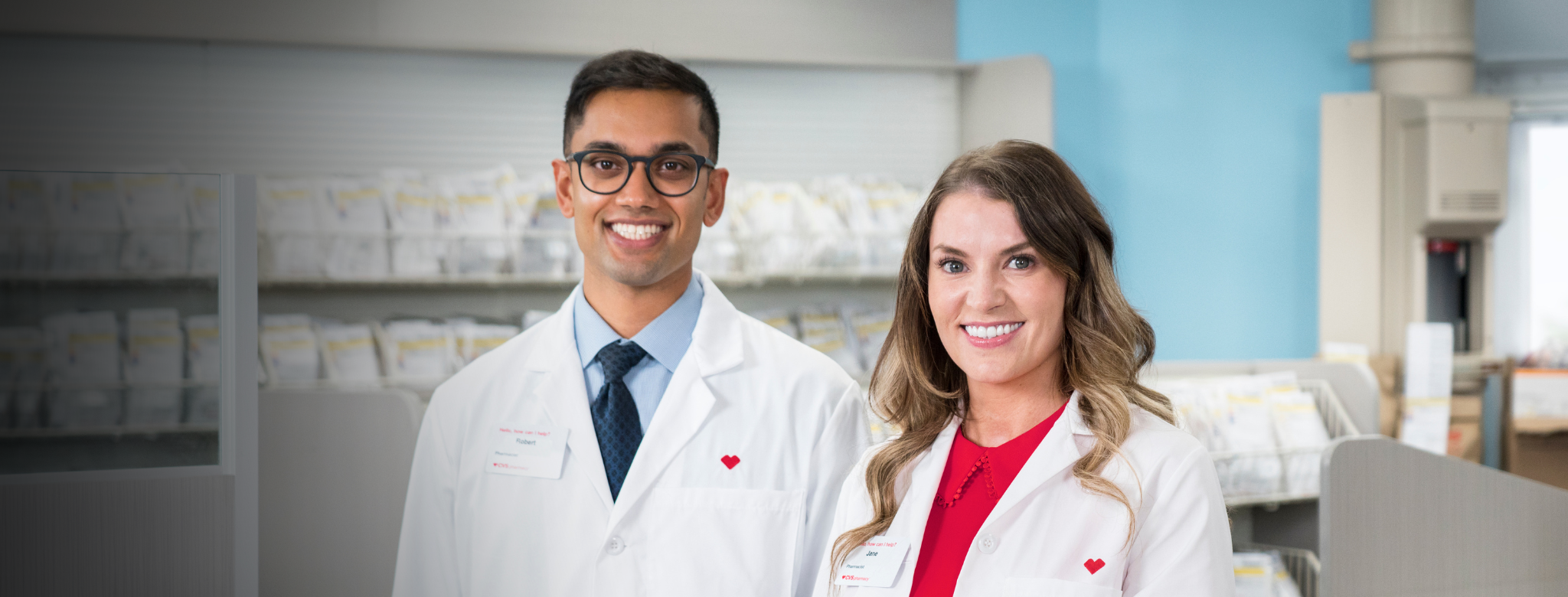 Two CVS Health pharmacy professionals in white lab coats stand in a pharmacy setting with prescription bins visible in the background. The individuals are facing the camera, and one is wearing a blue shirt and tie while the other is wearing a red top beneath the lab coat. The image highlights a professional pharmacy team environment.