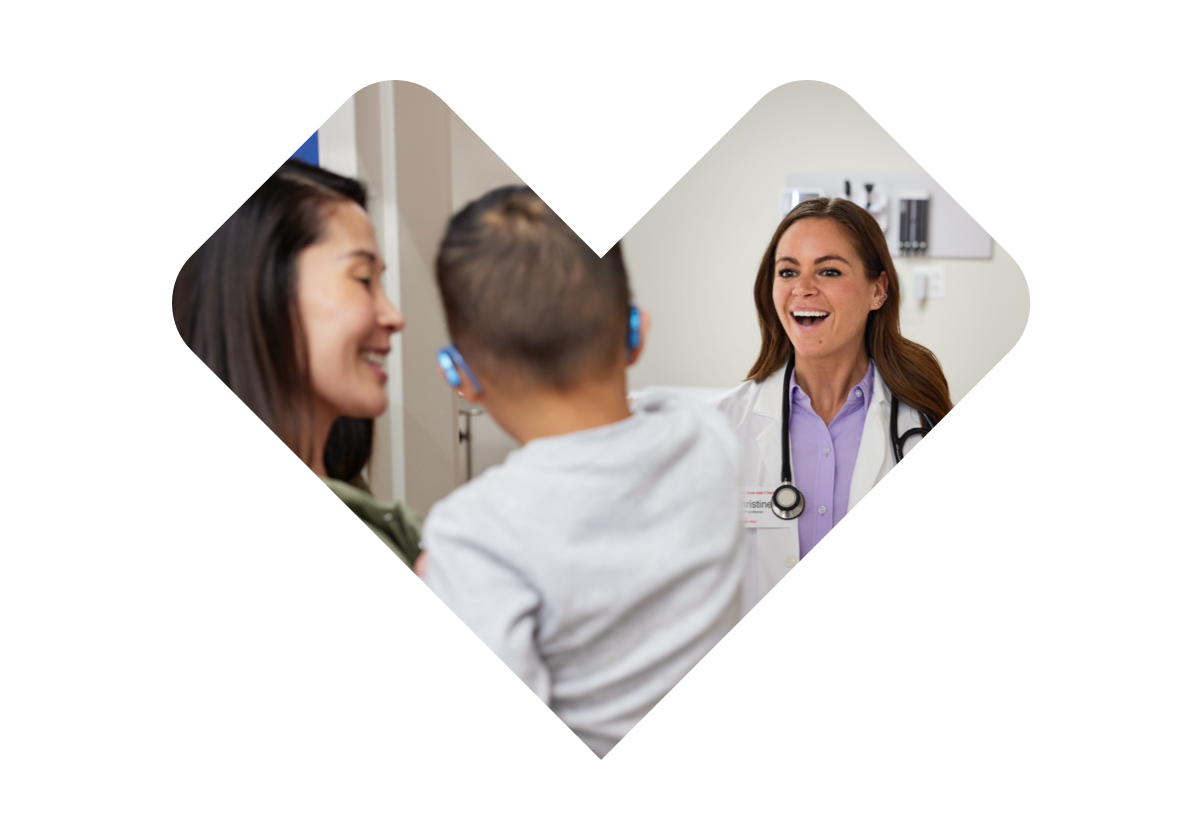 A doctor with a stethoscope smiles while speaking to a woman holding a young child. The child has hearing aids in both ears. Both the woman and doctor appear engaged and happy in the conversation within a clinical setting.