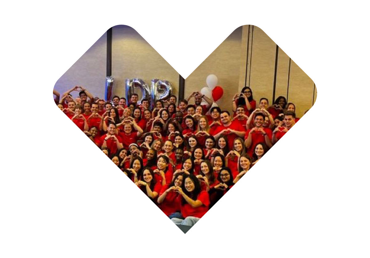 A large group of people wearing red shirts pose together for a photo in a heart-shaped frame. Some are making heart gestures with their hands. Balloons spell out "LDR" in the background. They appear to be in a celebratory or team-building event.