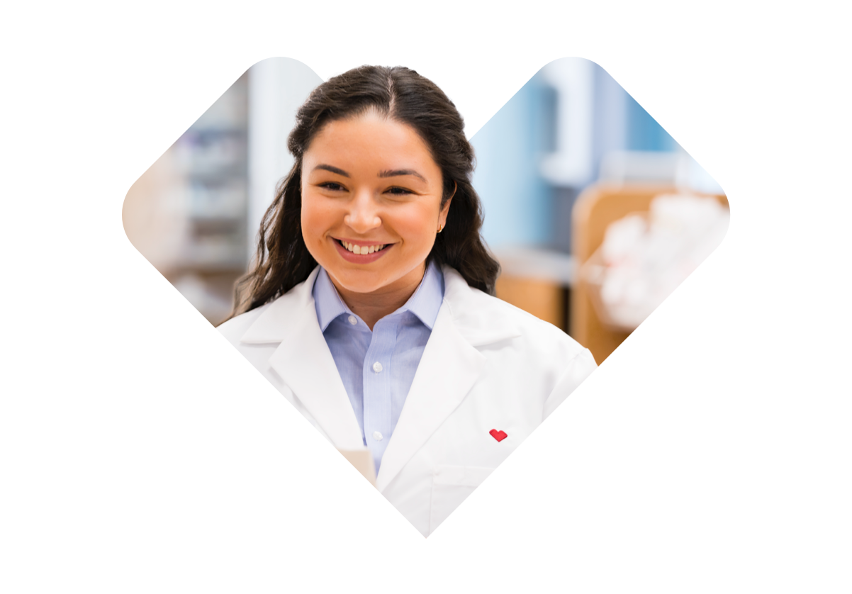 A smiling woman with long dark hair wearing a white lab coat and a light blue collared shirt stands in a bright room with blurred background. A small red heart is visible on her lab coat.