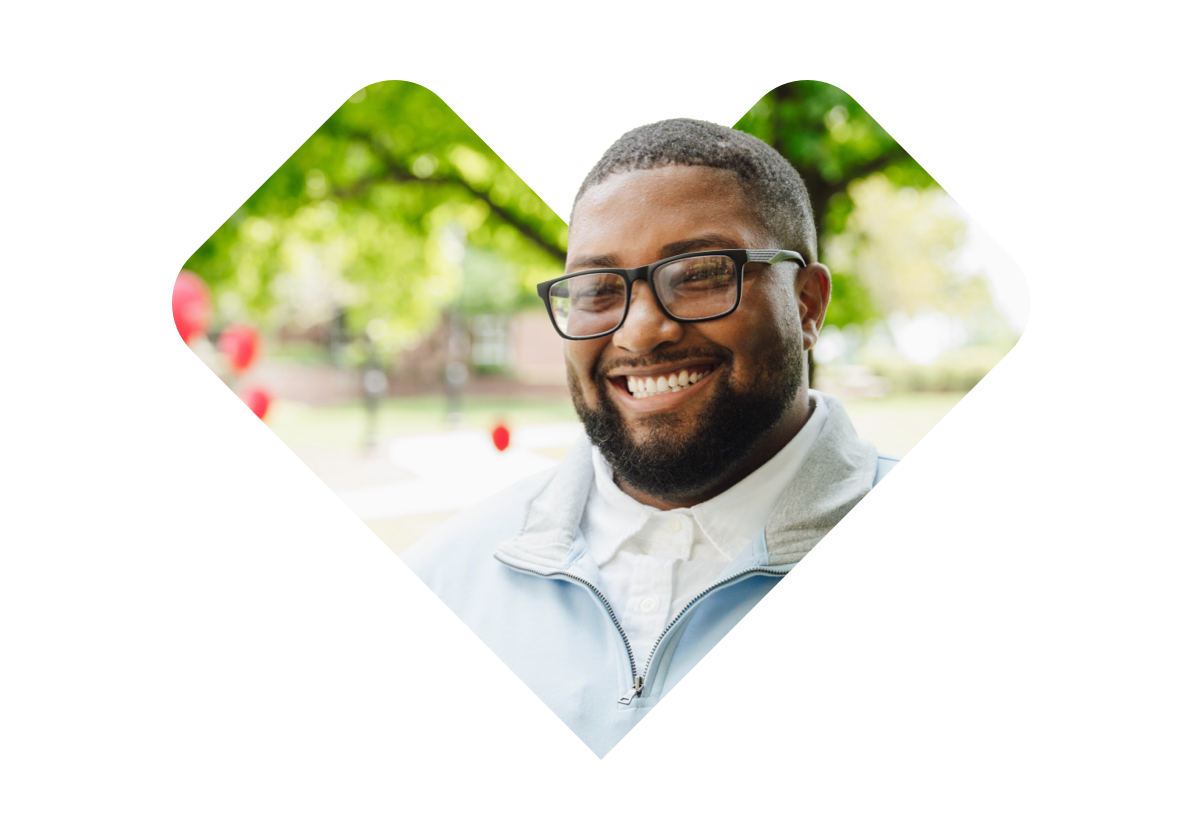 A smiling man with short black hair, a beard, and glasses is wearing a light blue jacket over a white shirt. He is outdoors, with blurred greenery and sunlight in the background, creating a colorful, diagonal abstract effect around the image.