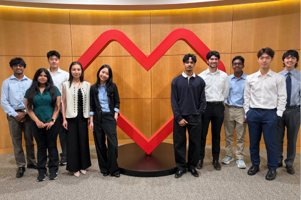 Group photo of ten interns standing in front of a large CVS red heart-shaped CVS Health logo mounted on a wood-paneled wall. The group is dressed in business and business-casual attire and arranged on both sides of the logo in a corporate office setting. Warm overhead lighting illuminates the space, and a gray speckled floor extends across the foreground.