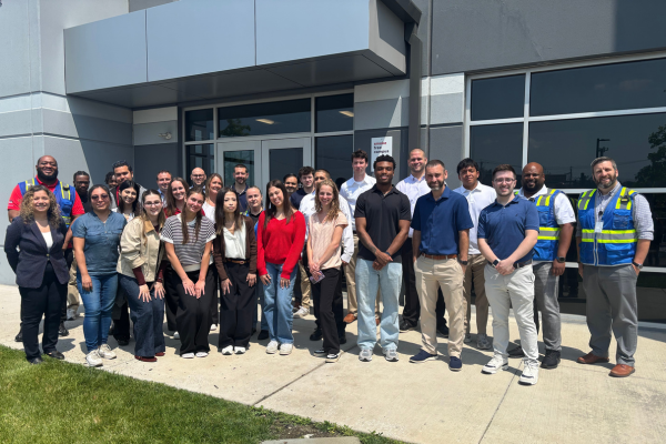 Group photo of approximately 30 people/interns standing outside the entrance of a modern commercial building. The group is gathered on a sidewalk in front of glass doors and large windows, with several individuals wearing business-casual attire and a few wearing high-visibility safety vests. Bright sunlight illuminates the scene, and a grassy area is visible in the foreground. The photo appears to capture a site visit or team gathering outside the facility.