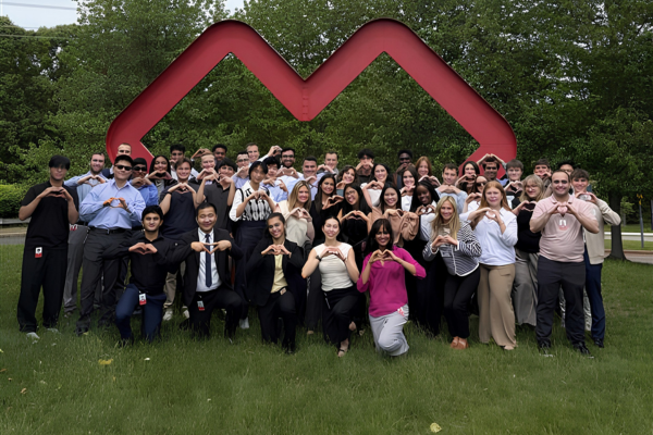 Large group photo of approximately 50 interns and employees gathered outdoors on a grassy lawn in front of a large red CVS Health heart logo sculpture. Many participants are forming heart shapes with their hands while posing for the photo. Trees surround the area in the background beneath an overcast sky, creating a campus-like setting. The group is dressed in a mix of business and business-casual attire and arranged in several rows around the logo.
