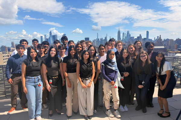 Group photo of approximately 25 interns standing on an outdoor rooftop terrace with a city skyline in the background. The group is arranged in several rows and dressed primarily in business-casual attire. The skyline features numerous high-rise buildings under a bright blue sky with scattered clouds. A paved terrace and metal railing frame the foreground, creating a professional and urban setting.