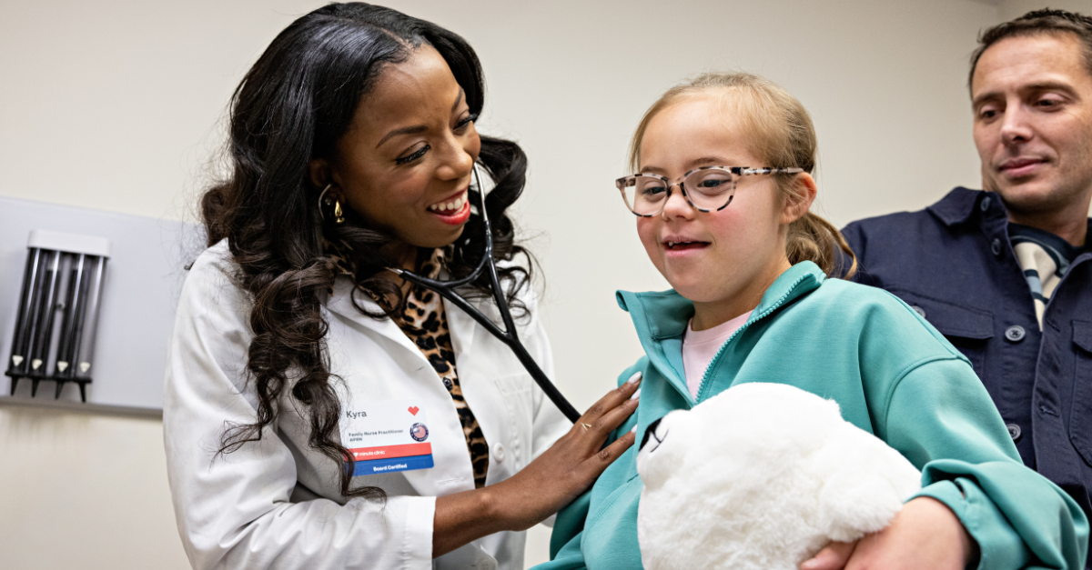 A healthcare provider in a white coat uses a stethoscope to listen to a young patient’s chest during an exam. The provider rests a supportive hand on the patient’s shoulder, while the patient holds a white stuffed animal. Another adult stands close by in the background.