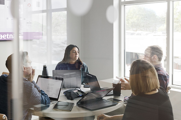 Four individuals are seated around a table in a well-lit conference room, engaged in a discussion. Laptops and notebooks are on the table, and they appear focused on the conversation. Outside, trees and cars are visible through the large windows.