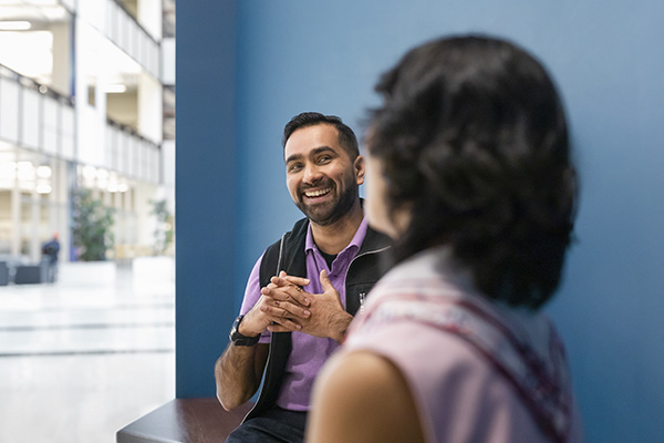 A man with a short beard and wearing a purple shirt and black vest is smiling and talking to a woman with shoulder-length dark hair, who is seen from the back. They are seated in an indoor space with blue walls and large windows.