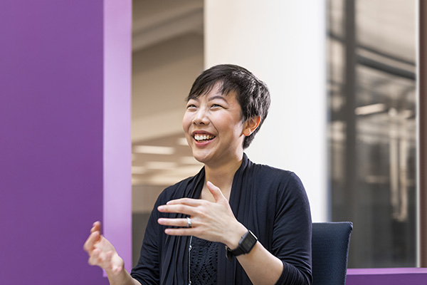 A person with short dark hair is sitting in a chair, gesturing with their hands, and smiling. They are wearing a black cardigan and a smartwatch on their left wrist. The background is purple and white with blurred office elements.