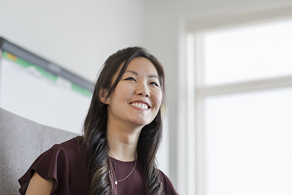 A woman with long, dark hair is smiling while sitting indoors. She is wearing a burgundy top and a simple necklace. In the background, part of a window and a blurry object, possibly a monitor, are visible. Bright natural light is coming through the window.