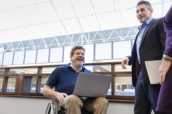 A man in a wheelchair is holding a laptop and smiling while talking to two colleagues, one holding a tablet. They are in a modern office setting with large windows and a high ceiling.