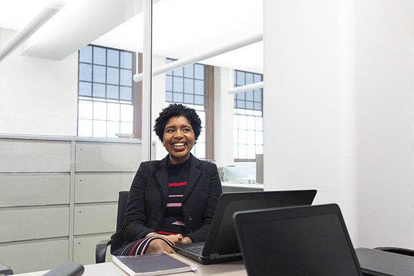 A person with short curly hair sits at a desk in an office with large windows. They are smiling and wearing a black jacket over a striped shirt. Two laptops and a notebook are on the desk in front of them.