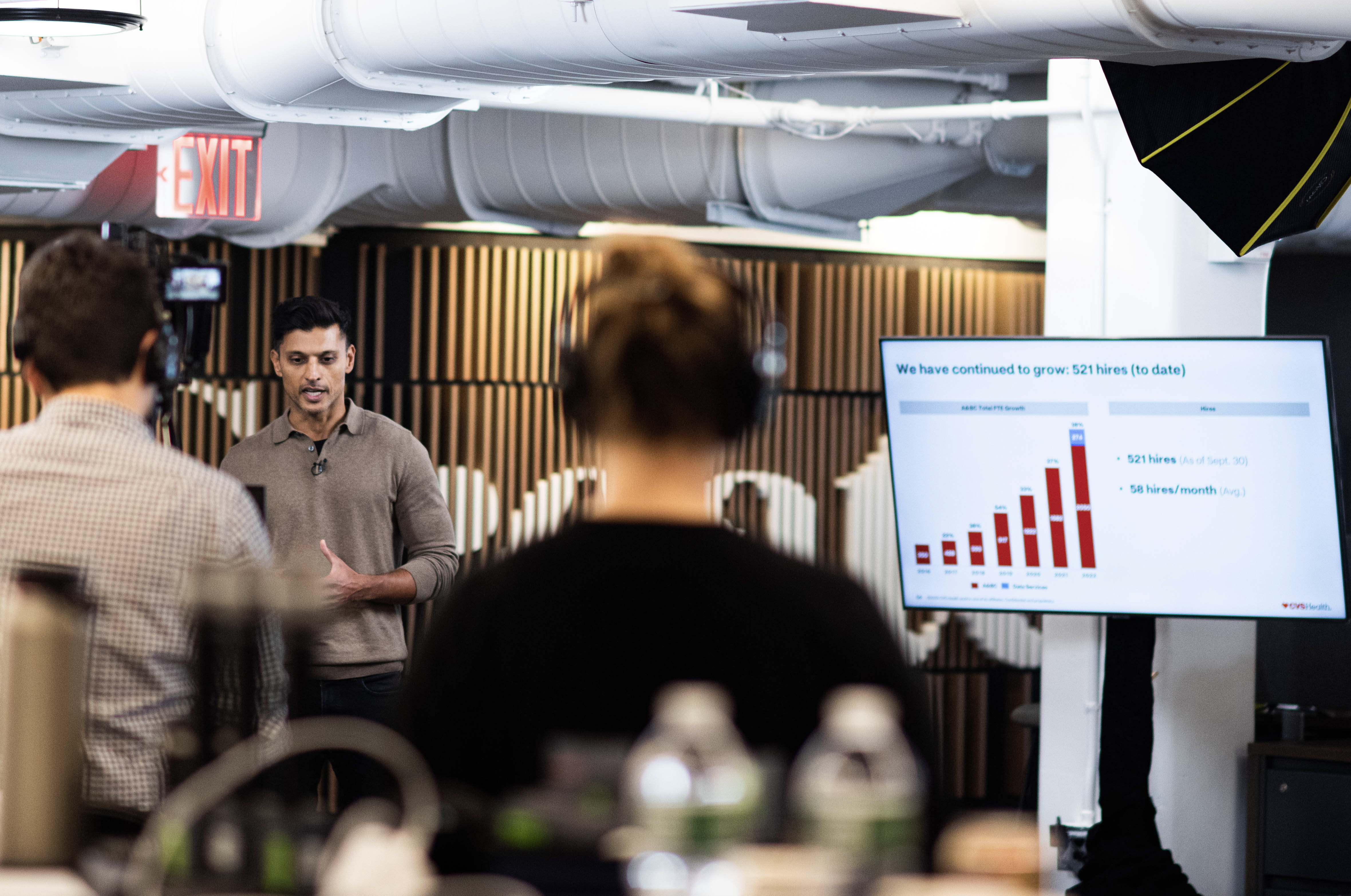 A person is delivering a presentation in a modern office setting. The person is speaking while a screen displays a bar graph showing growth statistics, with figures for K12 and NSF. Two people are seen in the foreground, one holding a camera and another wearing headphones.