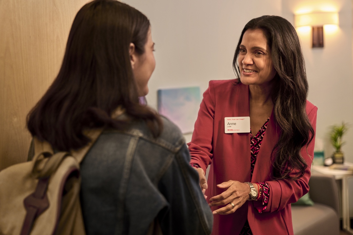 A woman with long hair in a red jacket, wearing a name tag, warmly greets a person with long dark hair wearing a denim jacket and backpack. They are indoors, in what appears to be an office or meeting room with soft lighting and decor in the background.