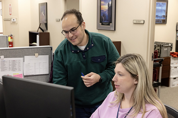 A man and a woman work together at a computer workstation in an office. The man, standing, points at something on the monitor while smiling. The woman, seated, looks attentively at the screen. Office cubicles and framed pictures on walls are visible in the background.