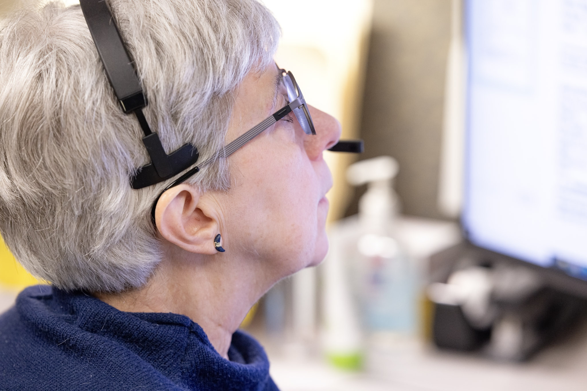 A person with short gray hair wearing a headset and glasses is attentively looking at a computer screen. The person is dressed in a dark blue top, and various office items, including a bottle of hand sanitizer, are visible in the blurry background.