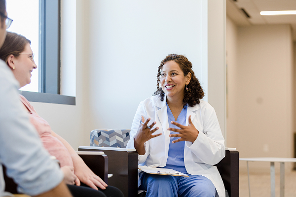 A woman wearing a white lab coat with a stethoscope draped around her neck stands in a medical office. She has a name badge that reads "Sarah." She is looking slightly to the side with a calm expression. The background includes an exam table and medical curtains.