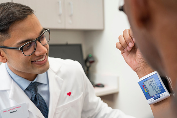 A healthcare professional in a white lab coat is smiling at a patient who is wearing a digital blood pressure monitor on their wrist. The monitor displays a reading of 120/80 with a pulse of 65. Medical equipment and a computer can be seen in the background.