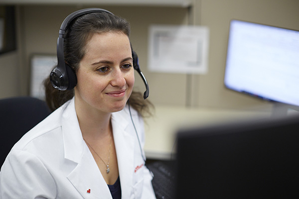 A woman wearing a white lab coat and a headset sits at a desk, looking at a computer monitor. She appears to be engaged in a conversation or conducting a telehealth session. The background includes office equipment and a blurred, large screen.