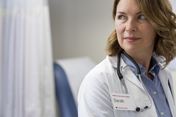 A woman wearing a white lab coat with a stethoscope draped around her neck stands in a medical office. She has a name badge that reads "Sarah." She is looking slightly to the side with a calm expression. The background includes an exam table and medical curtains.