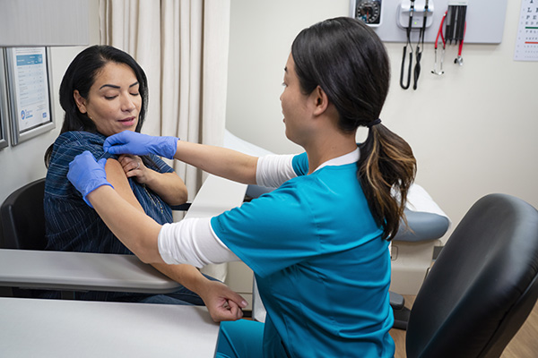 A healthcare professional wearing blue scrubs and gloves administers a vaccine to a patient. The patient is seated and pulling up her sleeve while the professional prepares the injection in a medical examination room equipped with medical instruments.